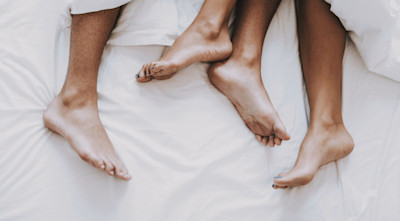 Unter einer Bettdecke schauen männliche und weibliche Füße hervor.