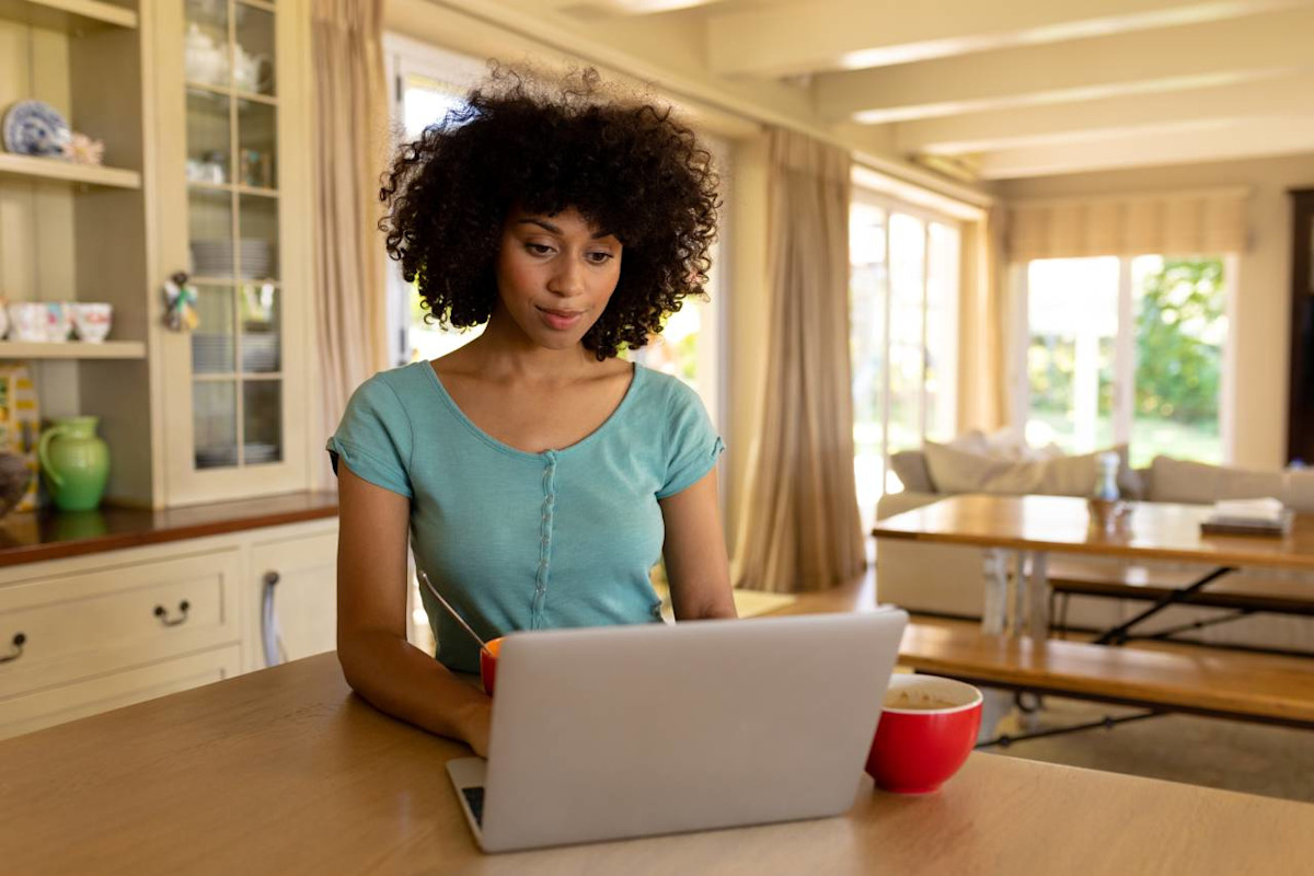 Junge Frau in blauen T-Shirt sitzt in der Küche vor ihrem Laptop.