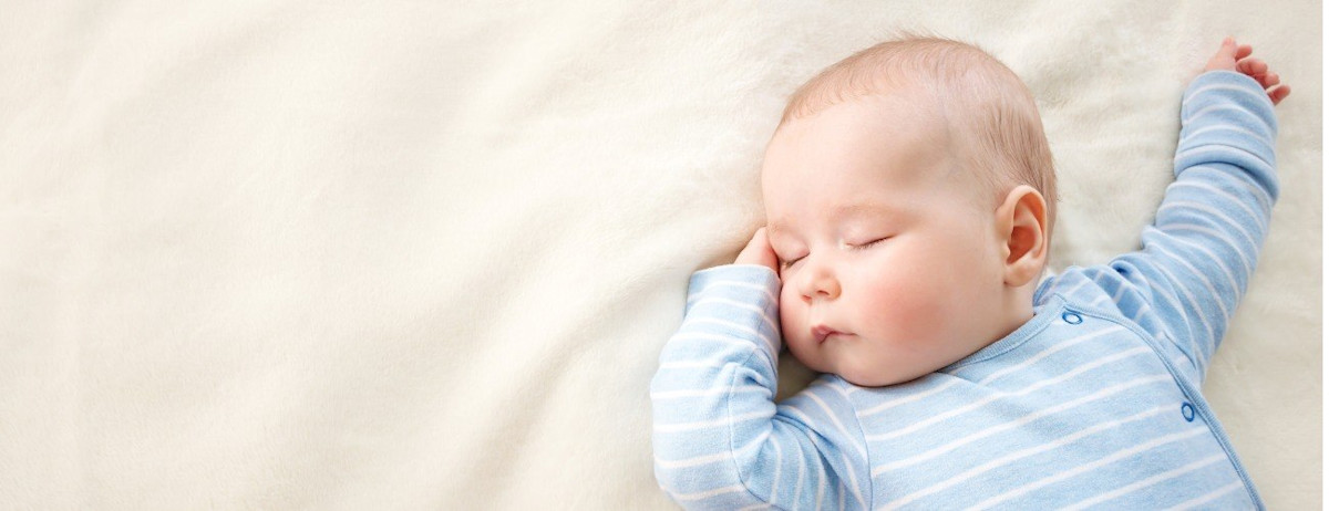 Baby mit blauem Strampler schläft auf heller Decke.