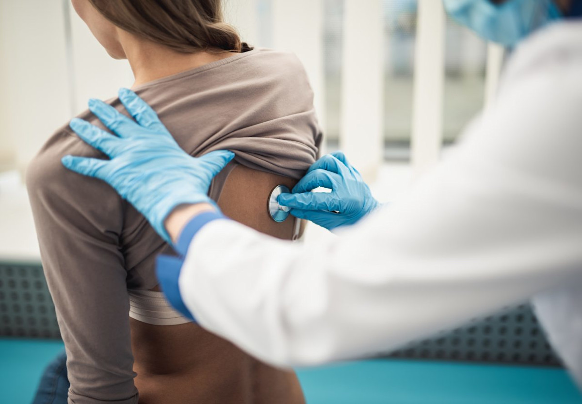 Ein Arzt hört mit dem Stethoskop am Rücken einer Patientin die Lunge ab.