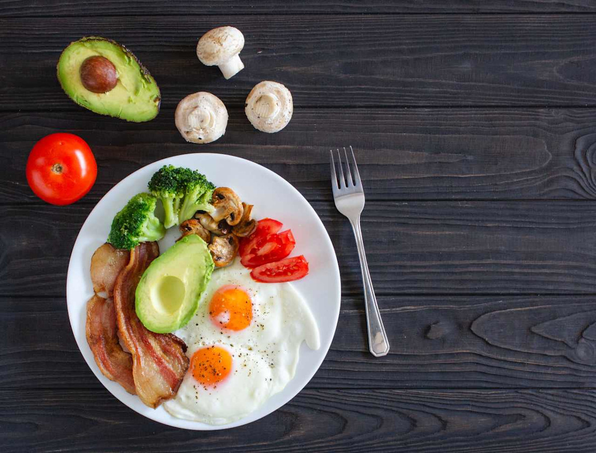 Es ist ein Teller mit ketogenen Lebensmitteln zu sehen, wie Avocado, Tomate, Pilzen, Broccoli, Speck und Ei.