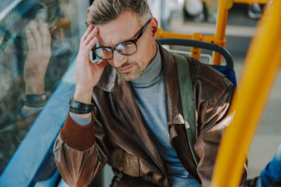 Ein Mann mit Brille sitzt im Bus und hält sich mit schmerzverzerrtem Gesicht den Kopf.