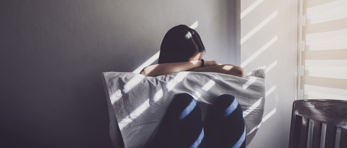 Junge Frau mit Migräne sitzt mit Kopfschmerzen und Übelkeit in einem abgedunkelten Raum.