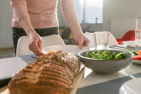 Im Bild ist ein gedeckter Tisch zu sehen, darauf aufgeschnittenes Brot und Broccoli. Eine Frau stellt einen Teller auf den Tisch.