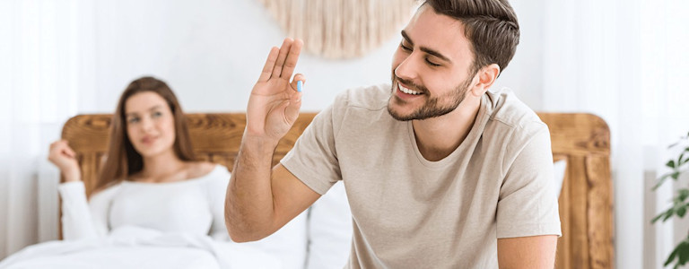 Mann sitzt auf dem Butt und hält eine blaue Tablette (Potenzmittel) hoch, Frau im Hintergrund.