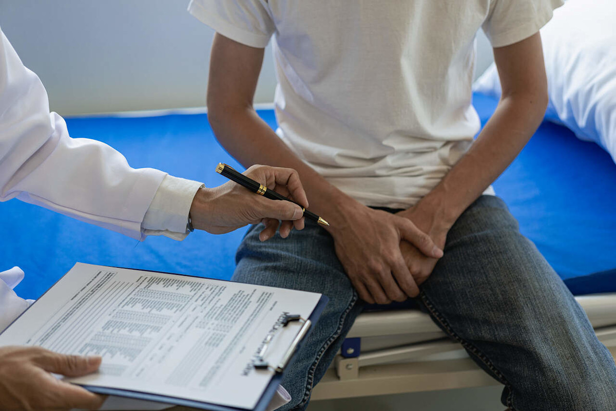 Ein Mann sitzt beim Arzt auf der Liege und hält sich vor Schmerzen den Intimbereich.