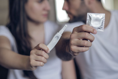 Eine Frau sitzt neben einem Mann. hält einen Schwangerschaftstest, er hält ein Kondom.