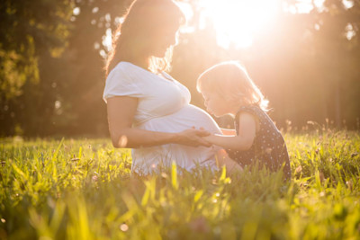 Schwangere Frau sitzt mit Mädchen auf einer Wiese in der Sonne.