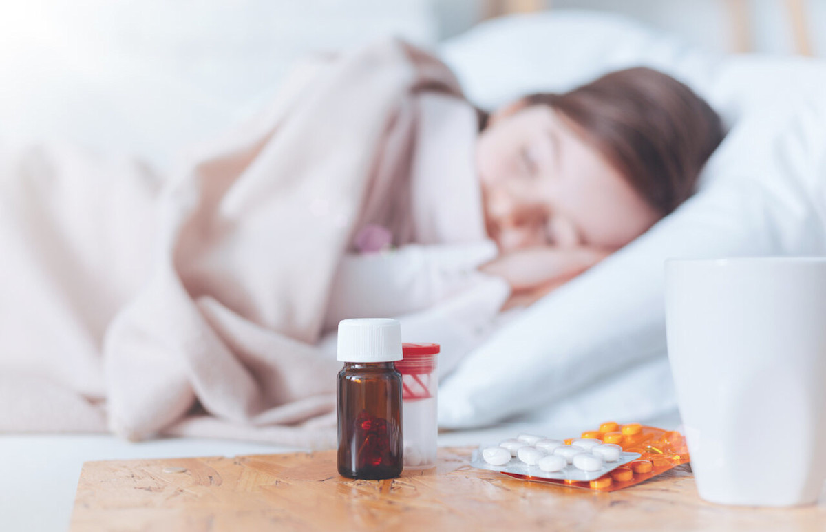 Schlaftabletten stehen auf einem Nachttisch, im Hintergrund schläft eine Frau im Bett.