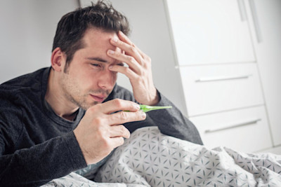 Ein Mann ist krank und sitzt im Bett. Er hat ein Fieberthermometer in der Hand.
