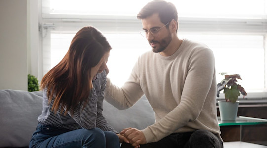 Mann sitzt mit besorgter Frau auf dem Sofa und Mann beruhigt sie