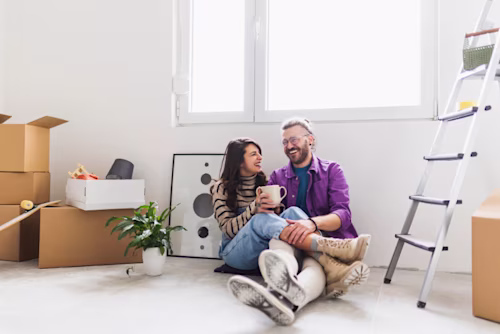 A couple sit on their living room floor surrounded by moving boxes, likely decorating their new house 
