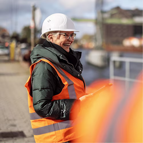 Woman worker with orange vest and white hardhat smiles at construction site