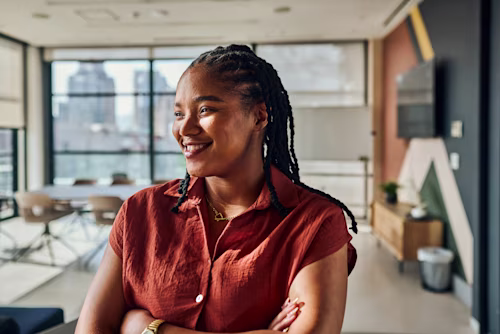 Retrato de una joven ingeniera de software negra sonriendo con los brazos cruzados, disfrutando de su trabajo en un entorno de oficina moderno.
