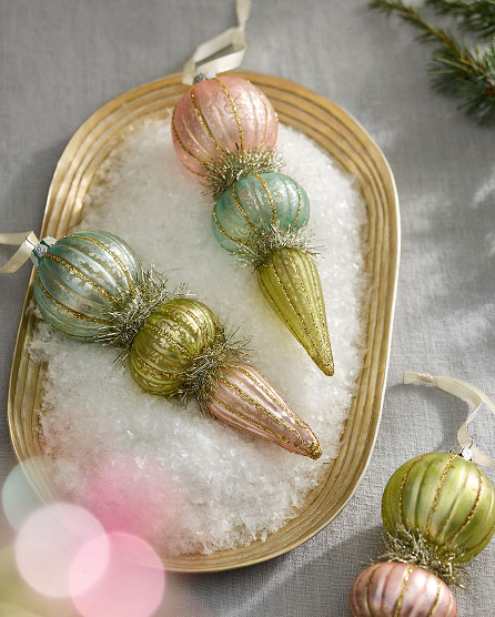 Pastel ornamental glass baubles in pink, green, and gold with glitter accents arranged on a wicker tray with white faux snow.