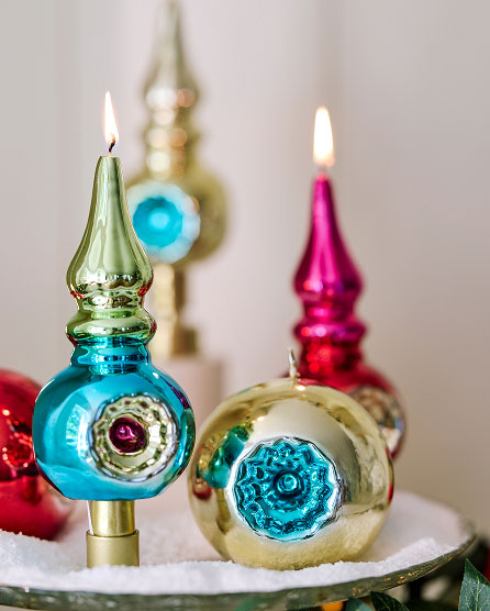 Colorful vintage-style glass Christmas tree toppers with lit candles in blue, pink, and gold finishes displayed on white surface.