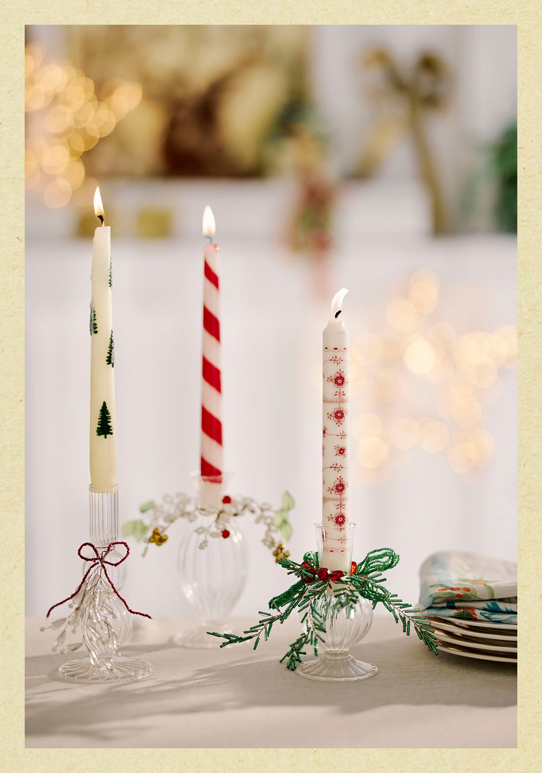 Three lit decorative Christmas candles in glass holders: white with trees, red and white striped, and white with snowflakes.