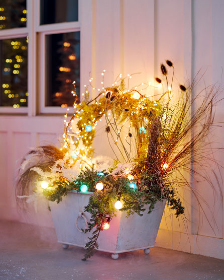 White planter filled with dried grasses, evergreen branches, and colorful string lights on a porch with pink door.