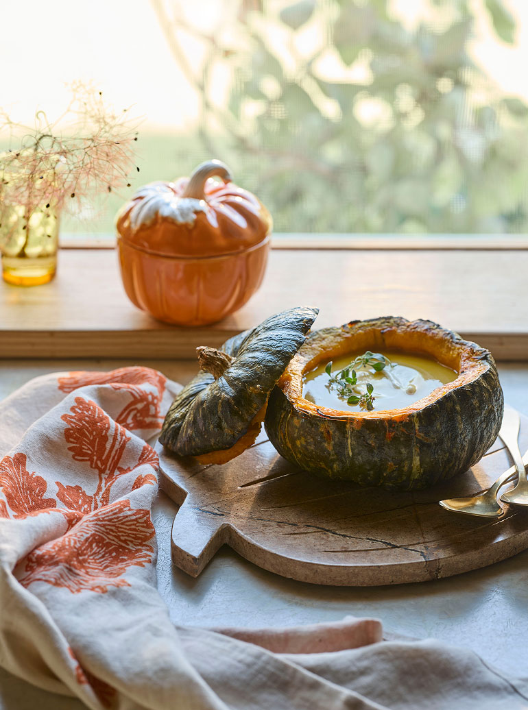 Creamy soup served in a hollowed green pumpkin on a wooden board with orange leaf towel and ceramic pumpkin container.