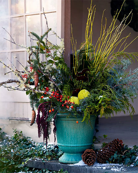Festive winter arrangement in turquoise urn with evergreens, red berries, yellow twigs, and pinecones on snowy doorstep.