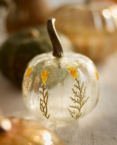 White decorative pumpkin with gold leaf accents and painted botanical design on a neutral surface with soft bokeh background.