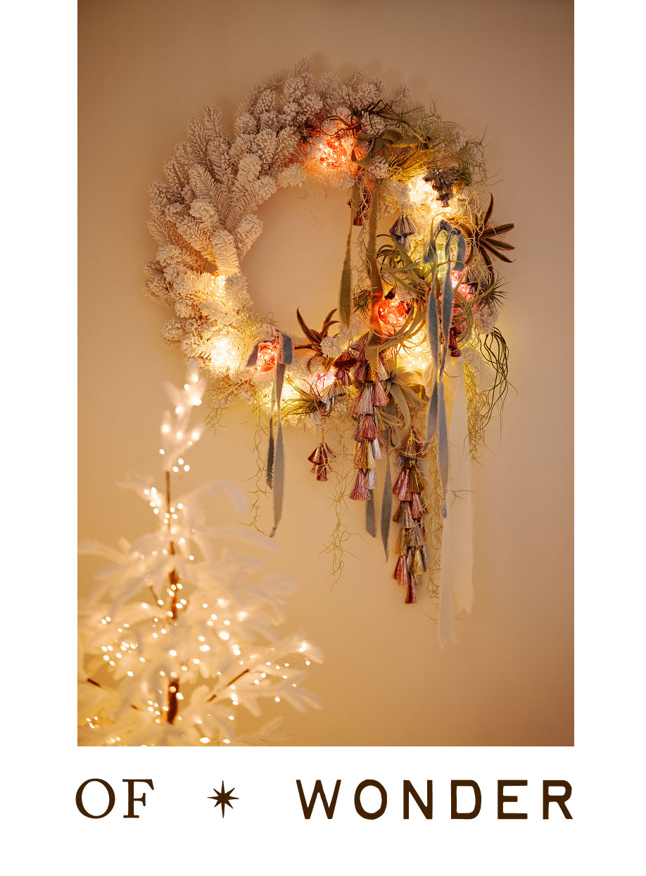 Illuminated white wreath with ornaments, antlers, and tassels on beige wall above lit white Christmas tree with text below.