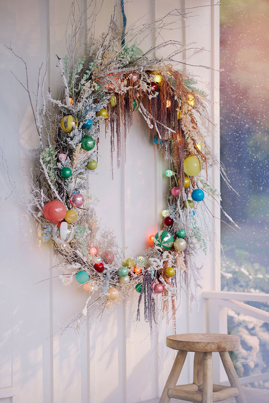 Rustic holiday wreath with colorful ornaments, frosted twigs, and draping tinsel hanging on white door with snowy window view.