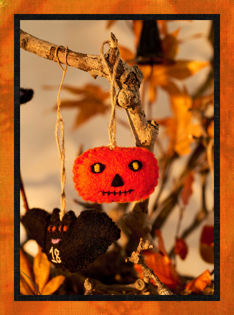 Orange felt pumpkin ornament with jack-o'-lantern face hanging from a twig branch with Halloween decorations in background