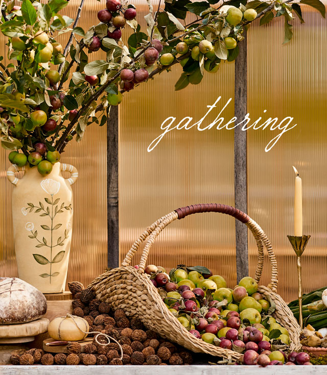 Rustic harvest display with fruits, bread, cheese, candles, and dried flowers on marble counter against textured glass wall.