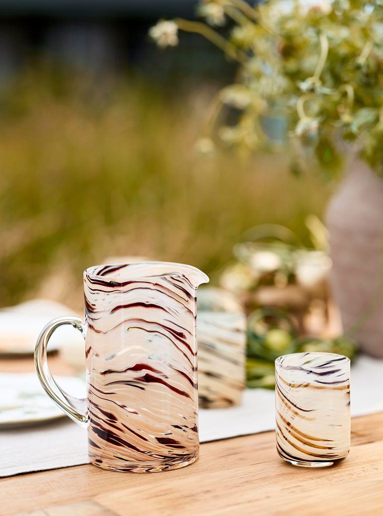 Glass pitcher and tumbler with swirled burgundy and amber patterns on a wooden table with blurred greenery in background.
