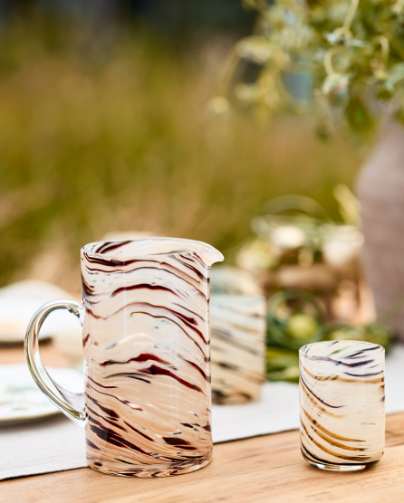 Glass pitcher and tumbler with burgundy swirl pattern on outdoor table with blurred greenery in background