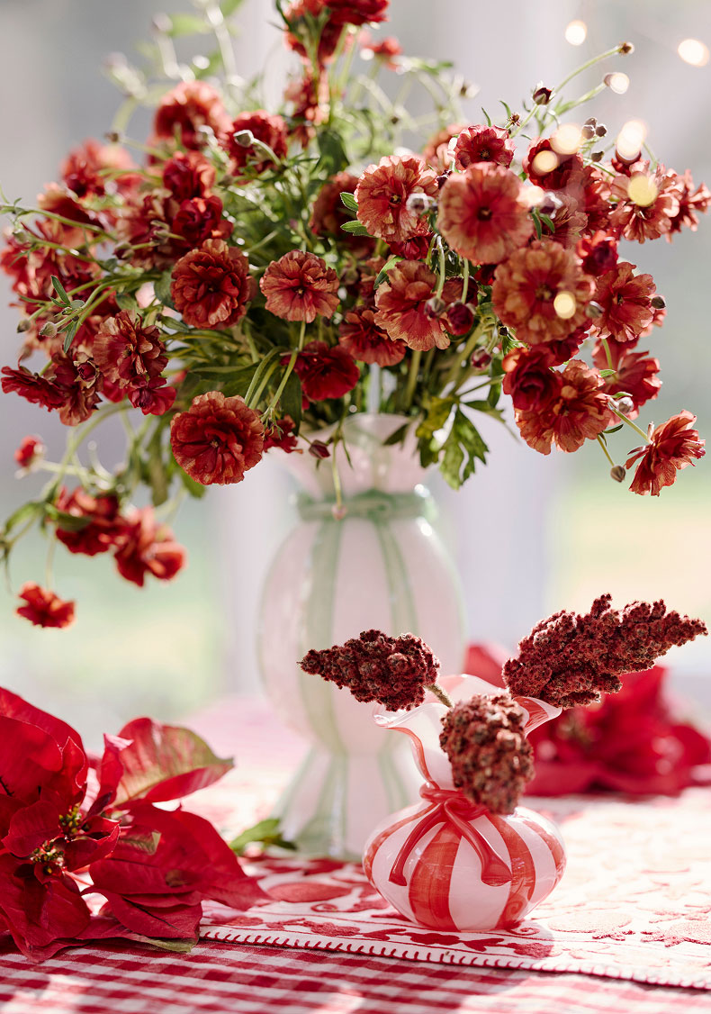 Red and coral flowers in a clear glass vase with holiday decorations on a festive red and white striped tablecloth.