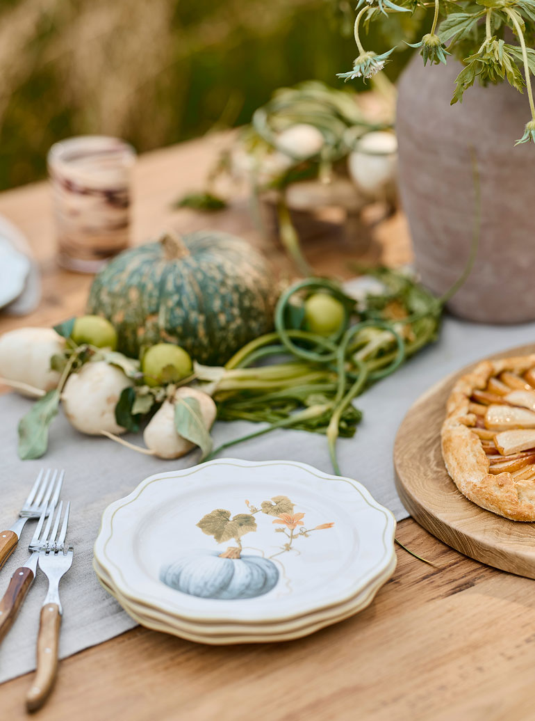 Autumn table setting with decorative pumpkins, gourds, white plates with pumpkin design, and a fruit tart on wooden board.