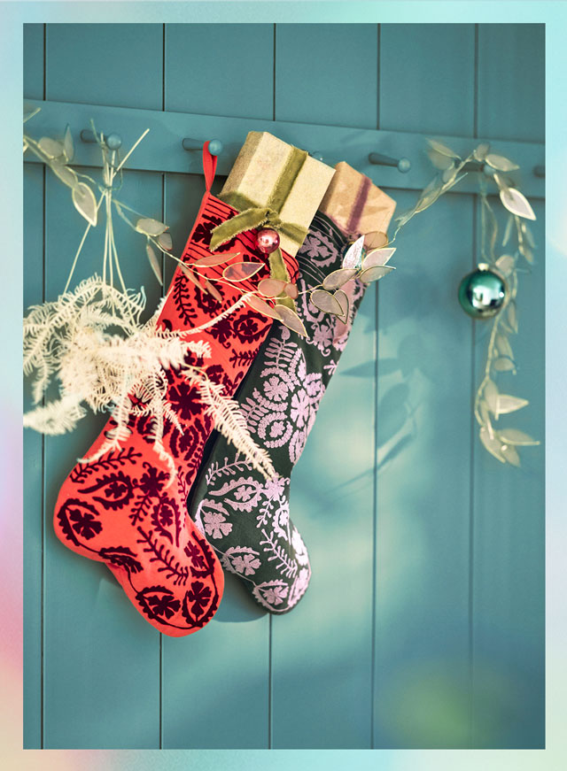 Two decorative Christmas stockings in red and dark green with floral patterns hang on a teal blue paneled wall with holiday decor.