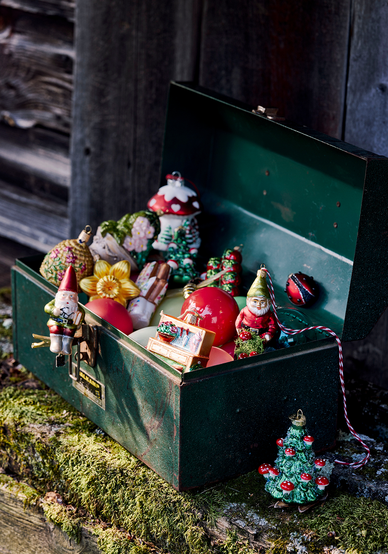Green metal toolbox filled with vintage Christmas ornaments including Santa figures, trees, and colorful decorations on mossy ground.
