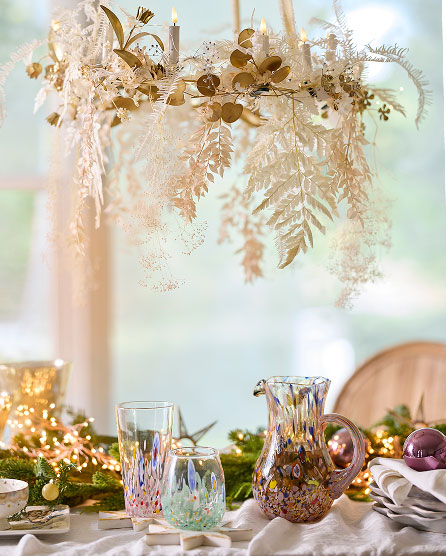 Elegant table setting with iridescent glassware, gold-accented greenery, and dried floral arrangement hanging overhead in cream and gold tones.