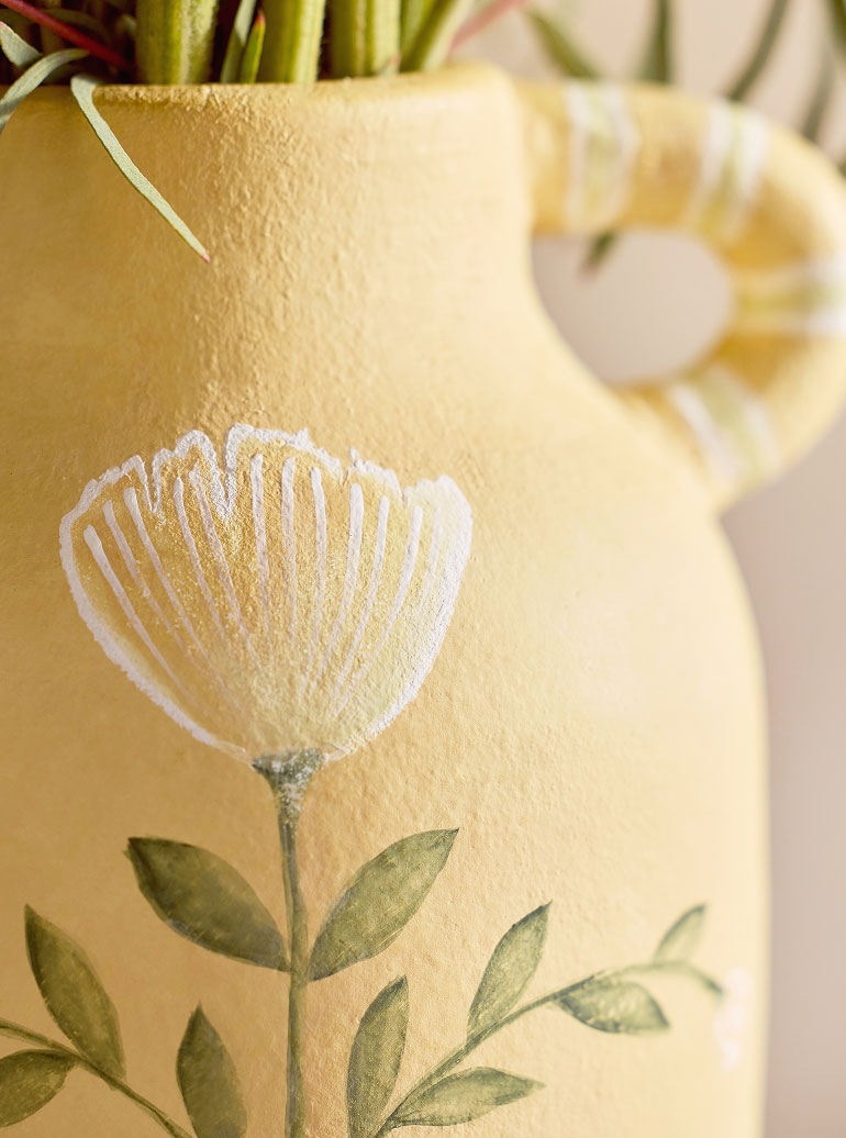 Yellow ceramic vase with white painted flower design and green leaves, with fresh stems visible at top.