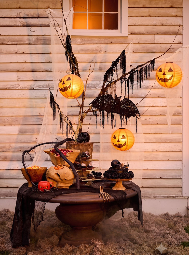 Halloween porch display with glowing jack-o'-lanterns, black decorative tree with bat and fringe, candy bowls, and skulls against white siding.