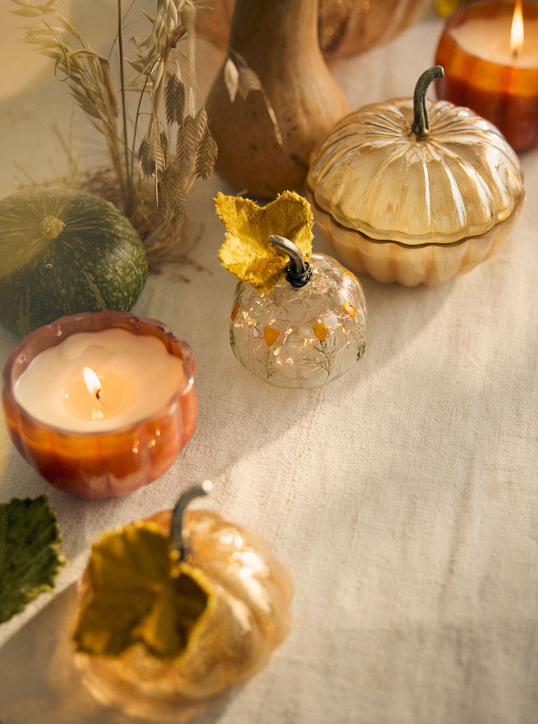 Autumn table display with decorative pumpkins, wheat stalks, glowing candles in amber holders, and fall leaves on cream linen.