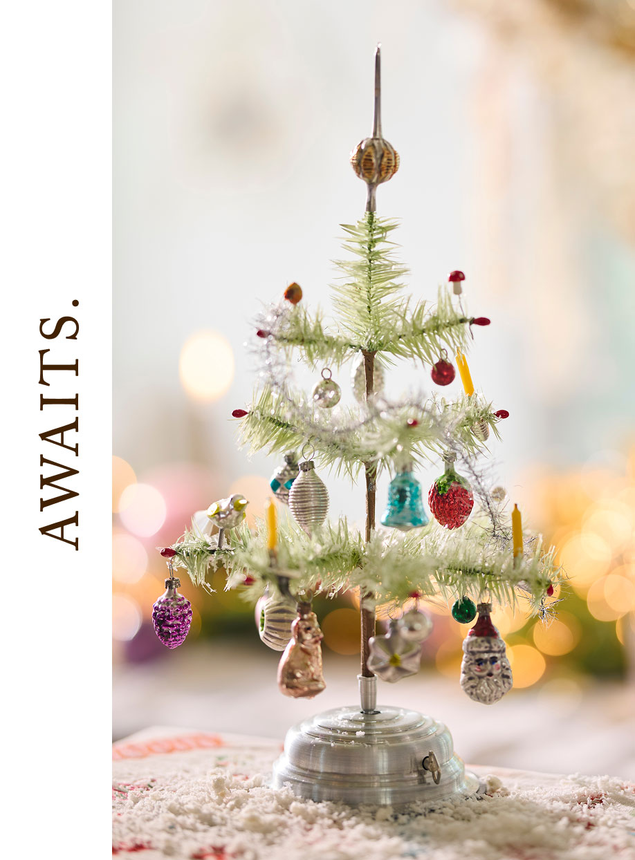 Small vintage-style Christmas tree with colorful glass ornaments, white garland, and candles on a metal stand.