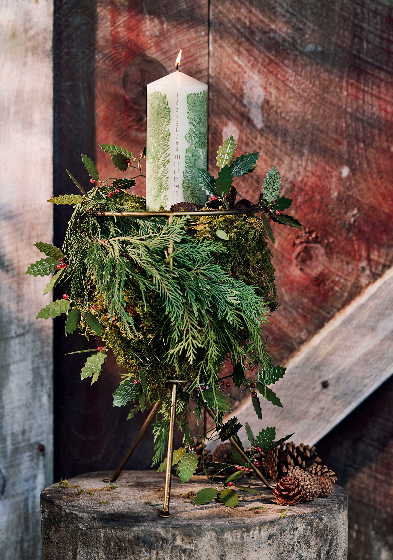 White pillar candle with green fern design sits atop evergreen wreath on gold stand with pinecones against rustic backdrop