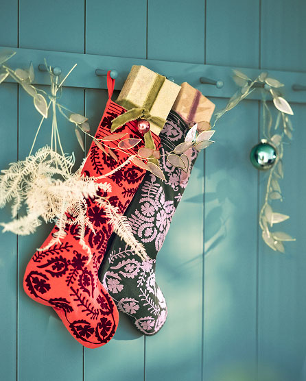 Two decorative Christmas stockings in red and black damask patterns hanging on a teal wall with white feather accents and gifts.