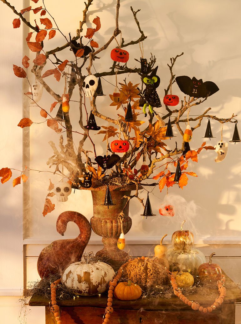 Halloween tree with orange autumn leaves, hanging pumpkin and skull ornaments, displayed in rustic urn with decorative pumpkins below.