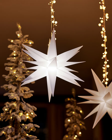 White illuminated star ornament hanging against warm golden lights with frosted pine decorations in background.