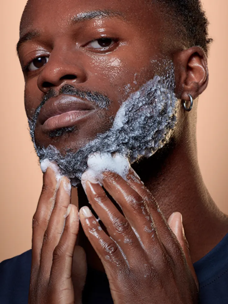Persona aplicando champú para barba con manos enjabonadas sobre fondo marrón.