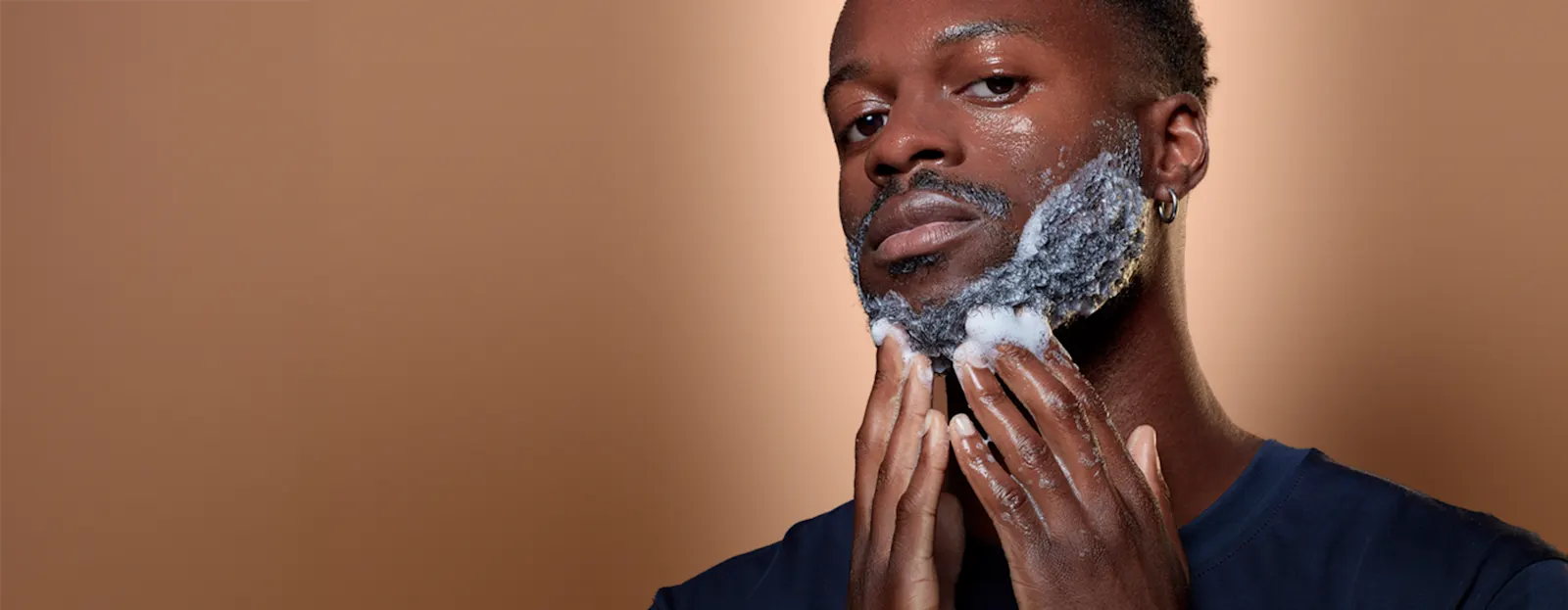 Persona aplicando champú para barba con manos enjabonadas sobre fondo marrón.