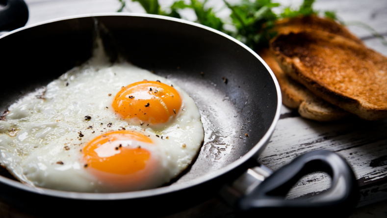 2 fried eggs in a non-stick pan with greens and toast in the background