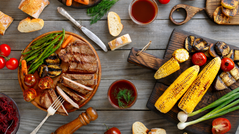 Grilled steak and vegetables on wooden boards, surrounded by condiments and bread, on a wooden background