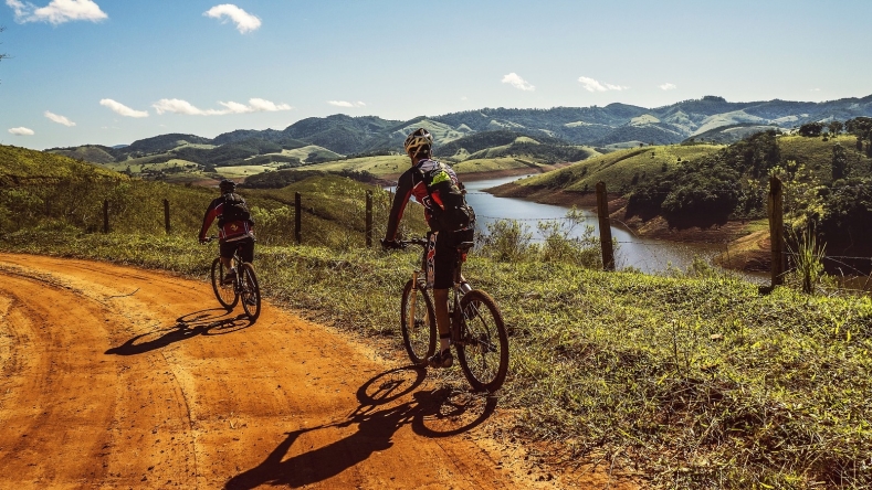 two people cycling along dirt road with mountains and water in the background