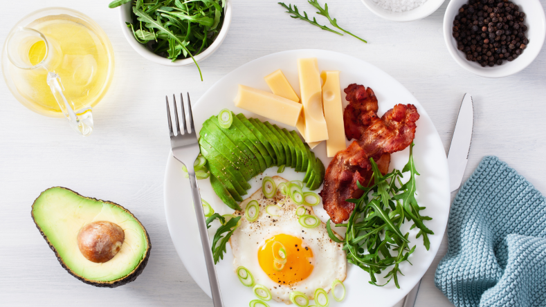 Ketogenic foods including bacon, greens, cheese and avocado, on a white plate on a white background